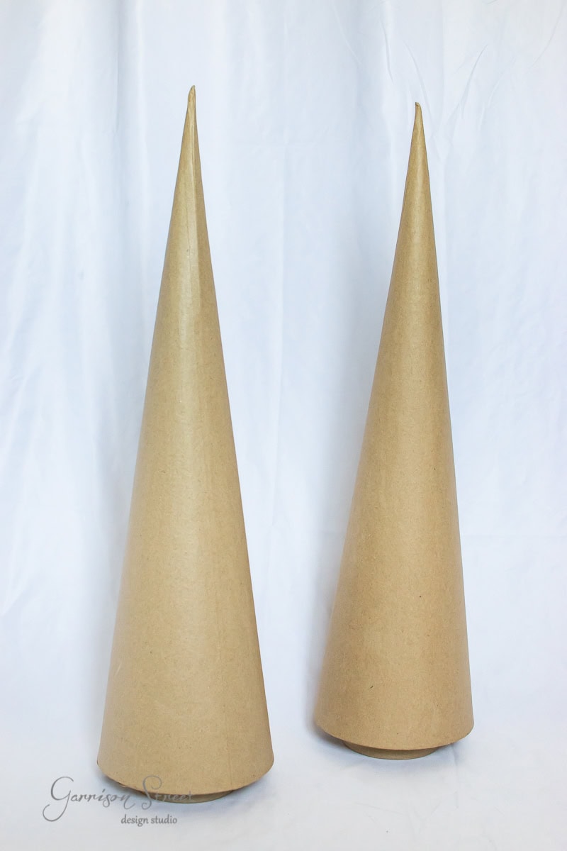 Two tall, cone-shaped, brown paper objects are standing upright against a white background, reminiscent of minimalist Christmas trees. The cones have a smooth surface and a pointed top, capturing the spirit of artisanal fottery design.