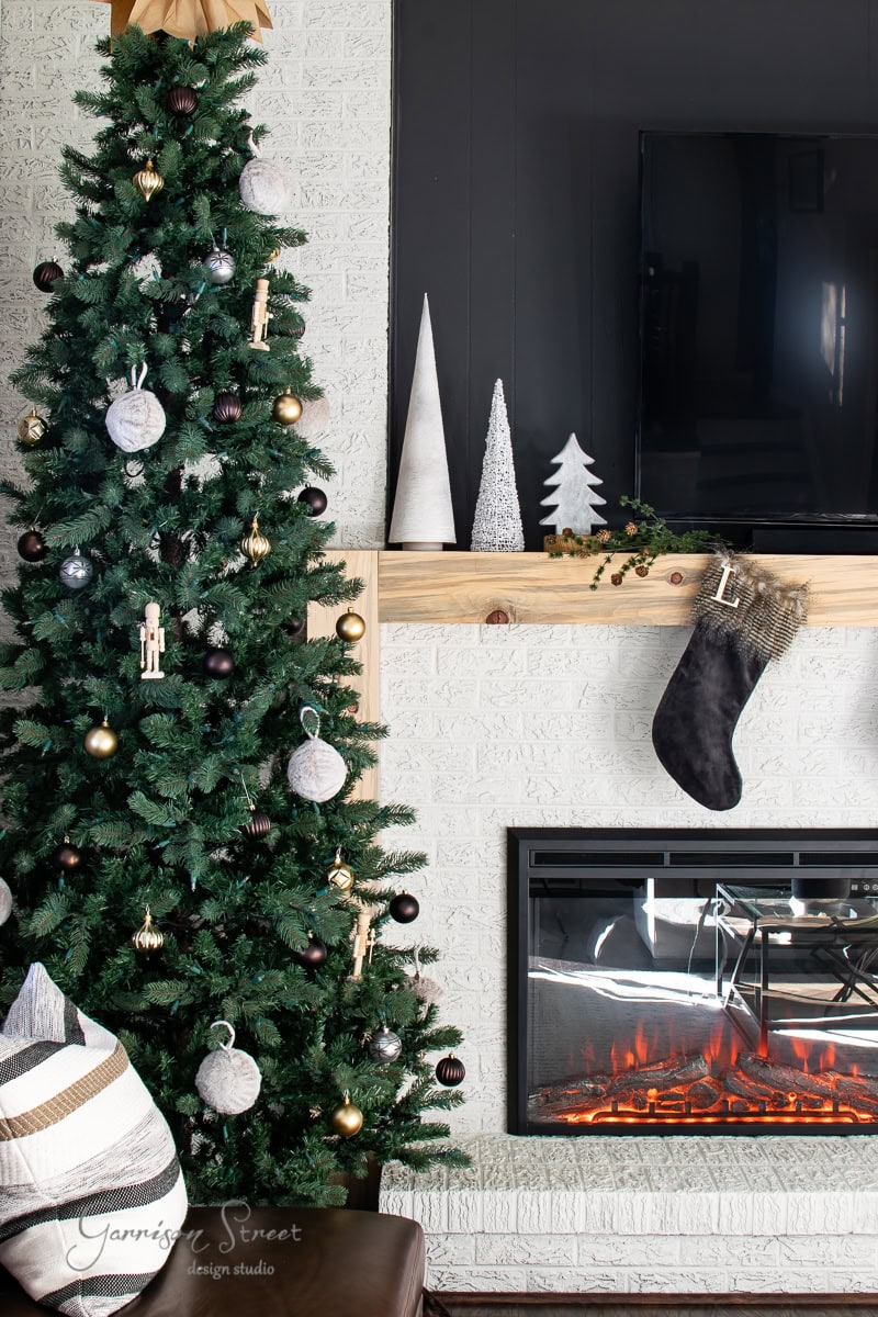 A decorated Christmas tree stands beside a fireplace. The mantel, an exquisite piece of fottery, is adorned with a stocking and miniature trees. A TV is mounted above the mantel, and the cozy scene is completed with a leather chair and pillows.