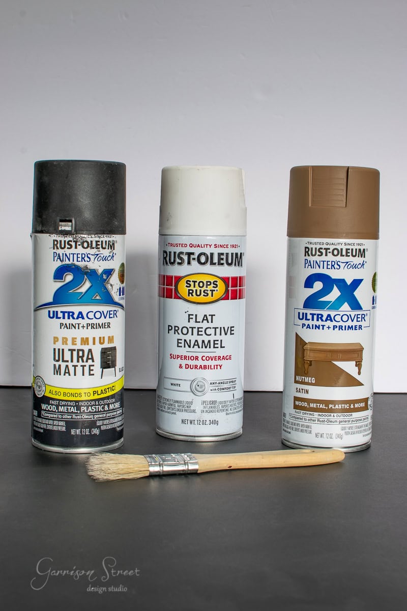 A photo of three Rust-Oleum spray paint cans and a paintbrush, perfect for crafting festive decorations like Christmas trees. The cans are labeled 2X Ultra Cover in black, white, and brown colors. The paintbrush rests in front on a flat surface.