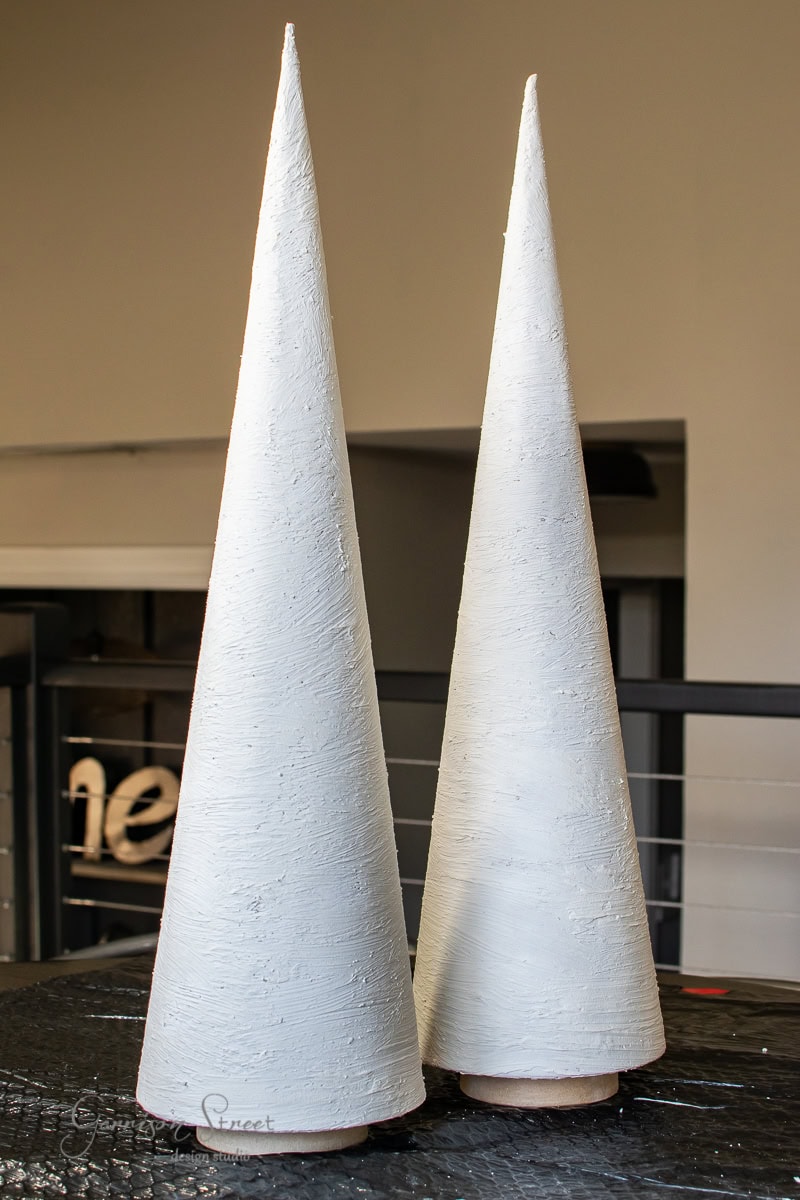 Two tall, white, cone-shaped sculptures with textured surfaces resemble elegant Christmas trees as they stand on a black surface. They are positioned in front of a blurred background featuring dark horizontal lines and part of a word.