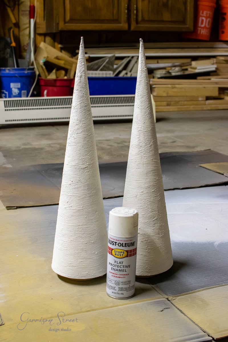 Two tall, white cone shapes resembling Christmas trees stand upright on a workshop floor, next to a can of Rust-Oleum flat protective enamel spray paint. The background features scattered wooden planks and tools, creating a scene reminiscent of festive pottery preparation.
