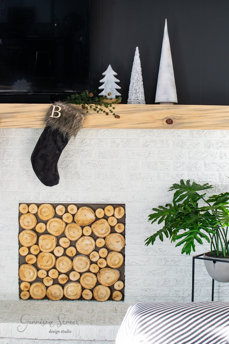 A cozy fireplace with a log pattern front, white brick surround, and wooden mantel. A black stocking with the letter 'B' hangs from the mantel, adorned with silver Christmas trees. Greenhouse plants are beside the fireplace, adding a touch of nature.