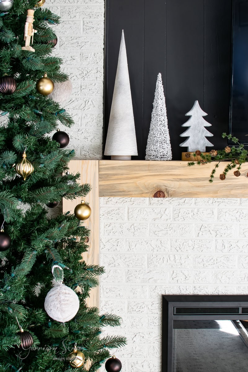 A decorated Christmas tree with gold and white ornaments stands beside a fireplace. On the wooden mantel are three pottery-inspired cone-shaped trees in white and silver, a small reindeer figure, and pine cones. The wall is painted white with a black accent.