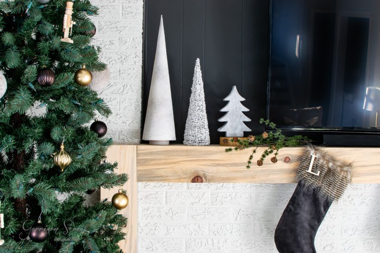 A cozy Christmas scene features a decorated tree with gold and brown ornaments on the left. The mantel beside it holds three decorative cones shaped like pottery trees, and a black stocking with a letter "L" dangles nearby. A TV sits to the right, enhancing the festive atmosphere.