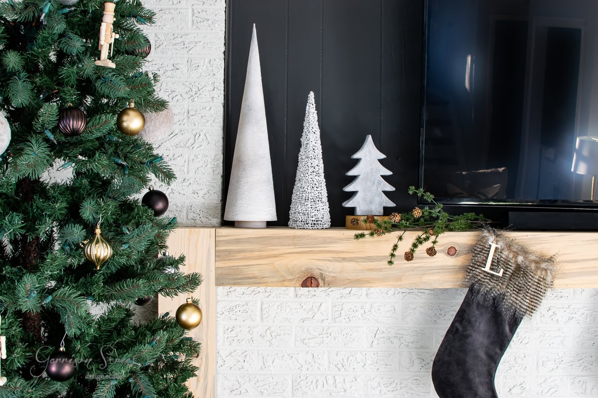 A cozy Christmas scene features a decorated tree with gold and brown ornaments on the left. The mantel beside it holds three decorative cones shaped like pottery trees, and a black stocking with a letter "L" dangles nearby. A TV sits to the right, enhancing the festive atmosphere.