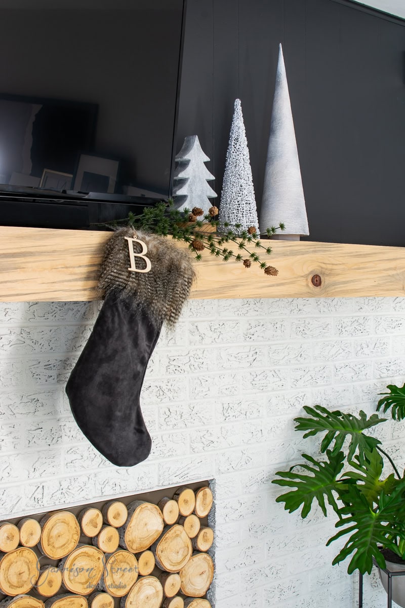 A black Christmas stocking with a letter "B" hangs from a wooden mantel decorated with pine branches and small pine cones. Above the mantel, three decorative cone-shaped Christmas trees add charm. Below, a section of stacked logs completes this cozy holiday scene.
