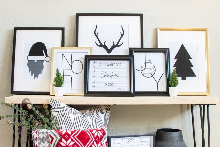 A shelf with framed Christmas-themed art, including a bearded figure with a Santa hat, antlers, a tree, and text like "Noel" and "Joy." The décor boasts small potted plants and seasonal cushions. Add charm to your space effortlessly with these printable holiday delights.