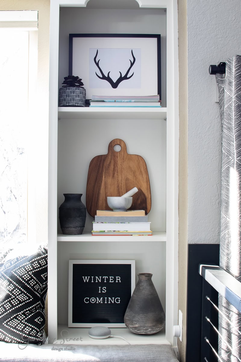 A white shelving unit with decorative items: framed antler print, patterned pottery, books, wooden cutting board, mortar and pestle, and a Christmas-themed letter board reading "WINTER IS COMING." Neutral tones with natural light from a nearby window. Perfect for displaying holiday printables.