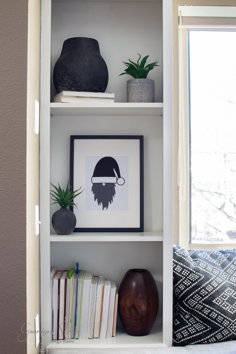A white bookshelf adorned with decorative items like two small green plants, black and gray pottery, a framed picture of a gnome, and a stack of books brings seasonal charm. Nearby, a window with a geometric-patterned cushion completes the cozy Christmas look.