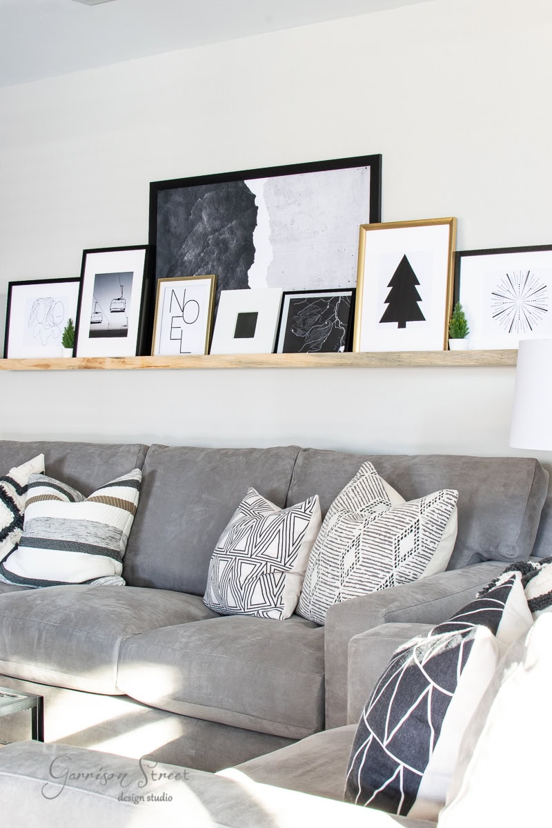 A modern living room features a gray sectional sofa adorned with patterned cushions. Above the sofa, two shelves display framed minimalist art, small decor pieces, and printable Christmas designs. The wall is a light neutral color, and sunlight brightens the festive space.