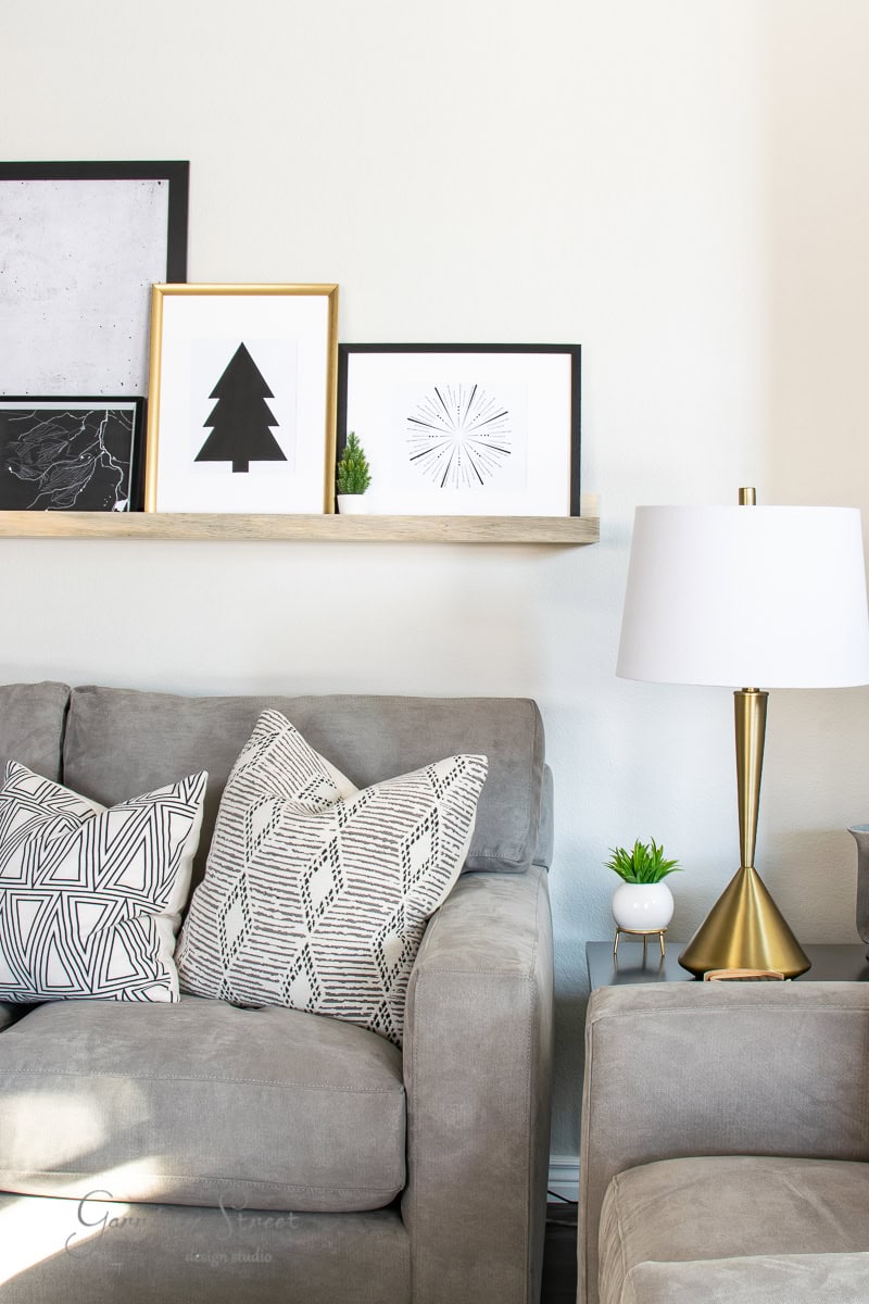 A cozy living room scene featuring a gray sofa with patterned pillows, a gold lamp on a side table, and a wooden shelf above displaying framed printable minimalist art and small plants against a white wall. Perfect for adding a festive touch this Christmas season.