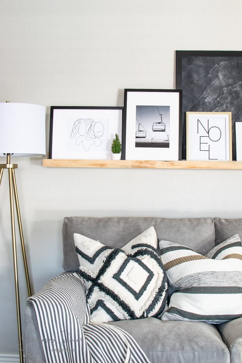 A cozy living room with a gray sofa adorned with black and white patterned pillows exudes festive charm. A wooden shelf displays framed artworks, including abstract and minimalist designs, alongside printable Christmas decorations. A floor lamp with a white shade stands beside the sofa.