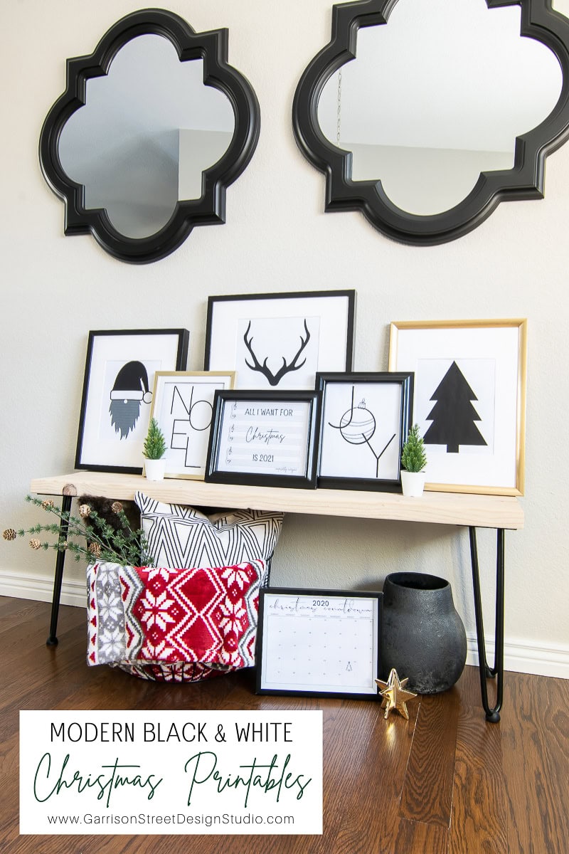 A display of black and white Christmas-themed framed art on a wooden bench with hairpin legs. Below, a basket contains red and white patterned blankets. Mirrors with decorative frames hang above, highlighting the "Modern Black & White Christmas Printables" available for download.