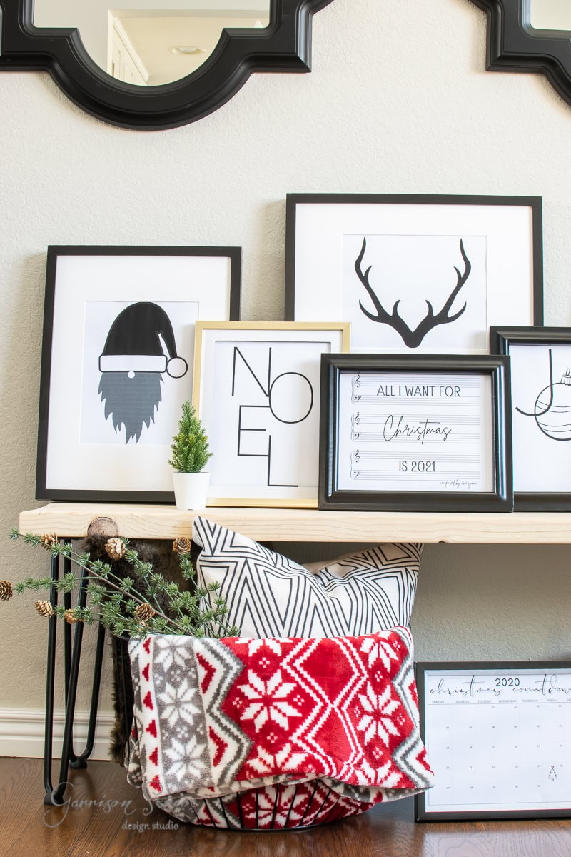 A holiday-themed display features printable framed prints: a Santa hat, "NOEL," antlers, and a wish list for Christmas 2021. Below, a basket holds a red and gray patterned blanket and geometric pillows. Christmas decor adds to the festive atmosphere.