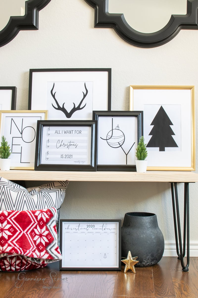 A shelf displays framed holiday-themed prints, including printable designs like deer antlers, a Christmas tree, and the words "Noel" and "Joy." Below, there's a red-patterned pillow, a small calendar, a star ornament, and a black vase with greenery.
