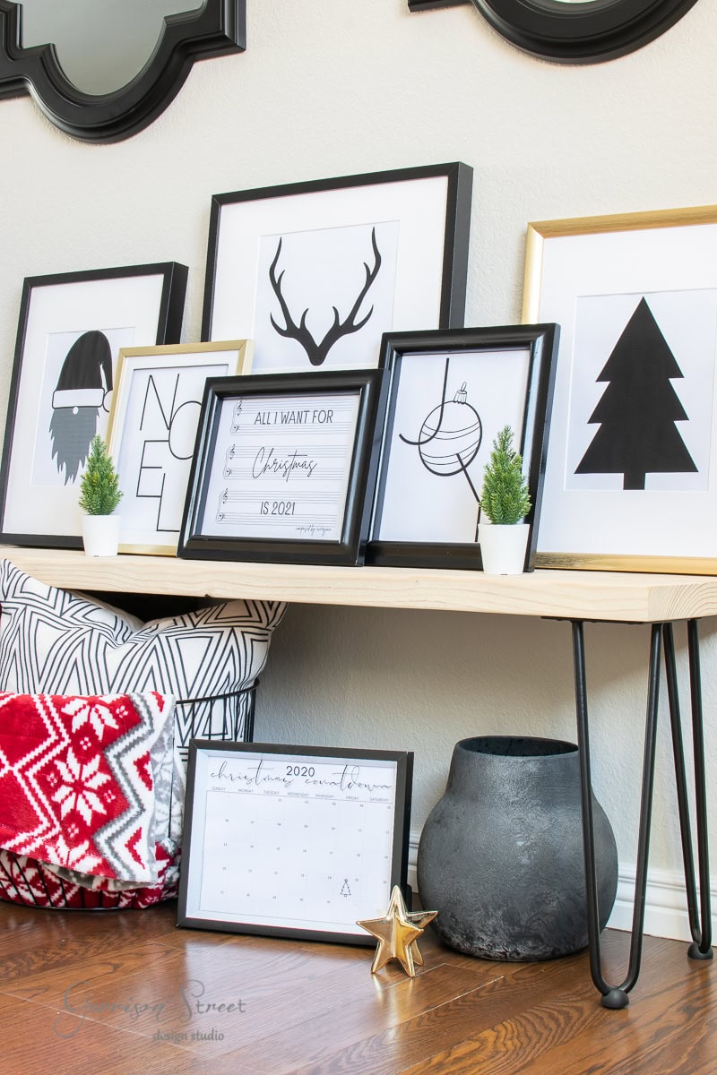 A minimalist Christmas-themed display on a wooden bench. Framed black and white art includes printable silhouettes of a reindeer, tree, and Santa. A 2020 calendar lies flat. Two small potted evergreens and a red patterned blanket add color.