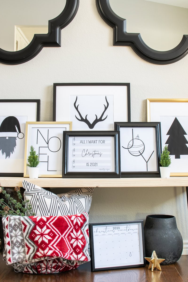 A decorative shelf displays framed Christmas-themed prints, including a reindeer, tree, and "Noel" sign. A red and white festive blanket is draped nearby. A small printable calendar marked December 2021 is visible, with a black vase and gold star accent.