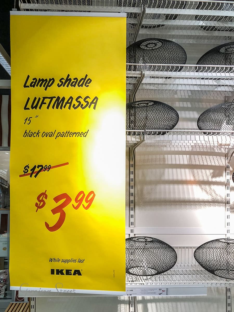 A large yellow IKEA sign showcases a "LUFTMASSA" black oval patterned lamp shade, once priced at $17.99, now just $3.99. These lamp shades rest on a white metal shelf, right beside the floating shelf display. Text at bottom reads "While supplies last".