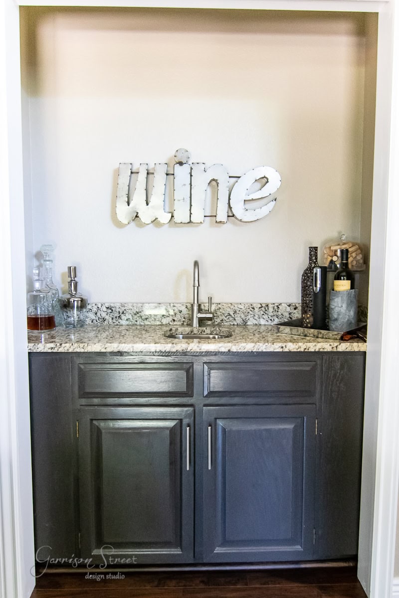 A small wet bar nook features dark cabinets, a granite countertop, and a sink. A floating shelf displays liquor bottles alongside a neatly arranged decanter, beneath a wall-mounted sign that reads "wine.