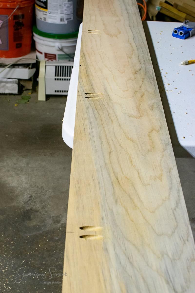 A long wooden board, destined to become a floating shelf, rests on a table in the workshop. Its edge features multiple pocket holes. Tools and supplies, including a drill and containers, are visible amidst the slightly wet background.