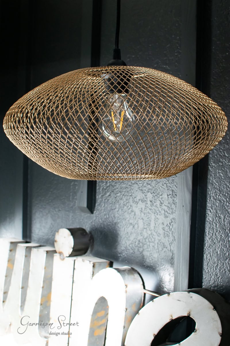 A modern pendant light with a gold wire mesh design hangs in front of a dark textured wall, casting subtle illumination over the wet bar. A clear bulb is visible inside the mesh, while below, partial metal letters adorn a floating shelf.