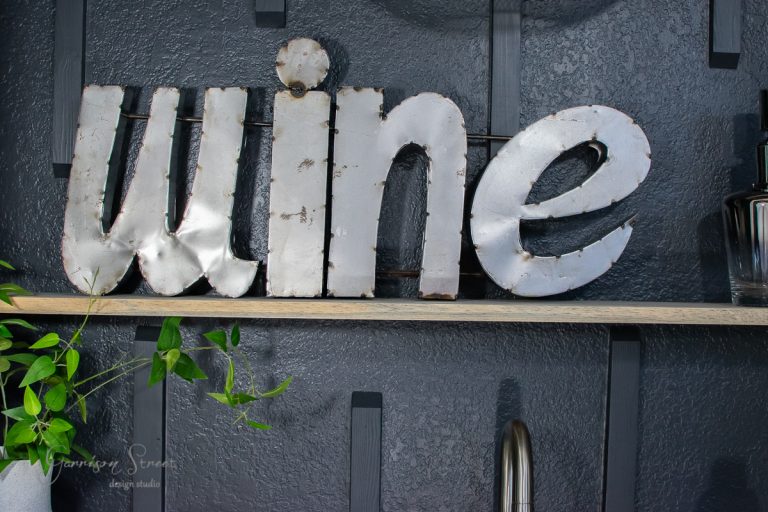 Metal letters spell "wine" on a dark gray wall. They're mounted on a floating wooden shelf next to a small plant with green leaves. This inviting setup adds a charming touch to your home bar area, seamlessly merging elegance with functionality.