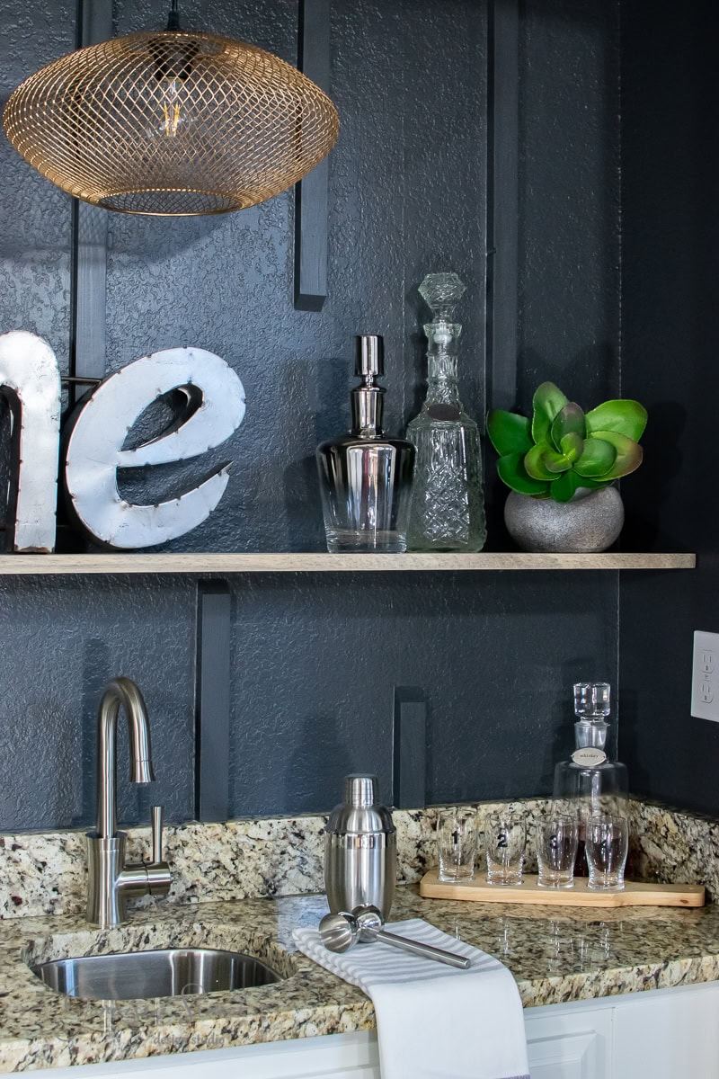 A modern wet bar kitchenette with dark walls and a granite countertop features a metal faucet, cocktail shaker, glass decanters, and a green potted plant. A floating shelf showcases elegant decor beneath the wire-frame light fixture, while a towel drapes over the sink edge next to the decorative sign.