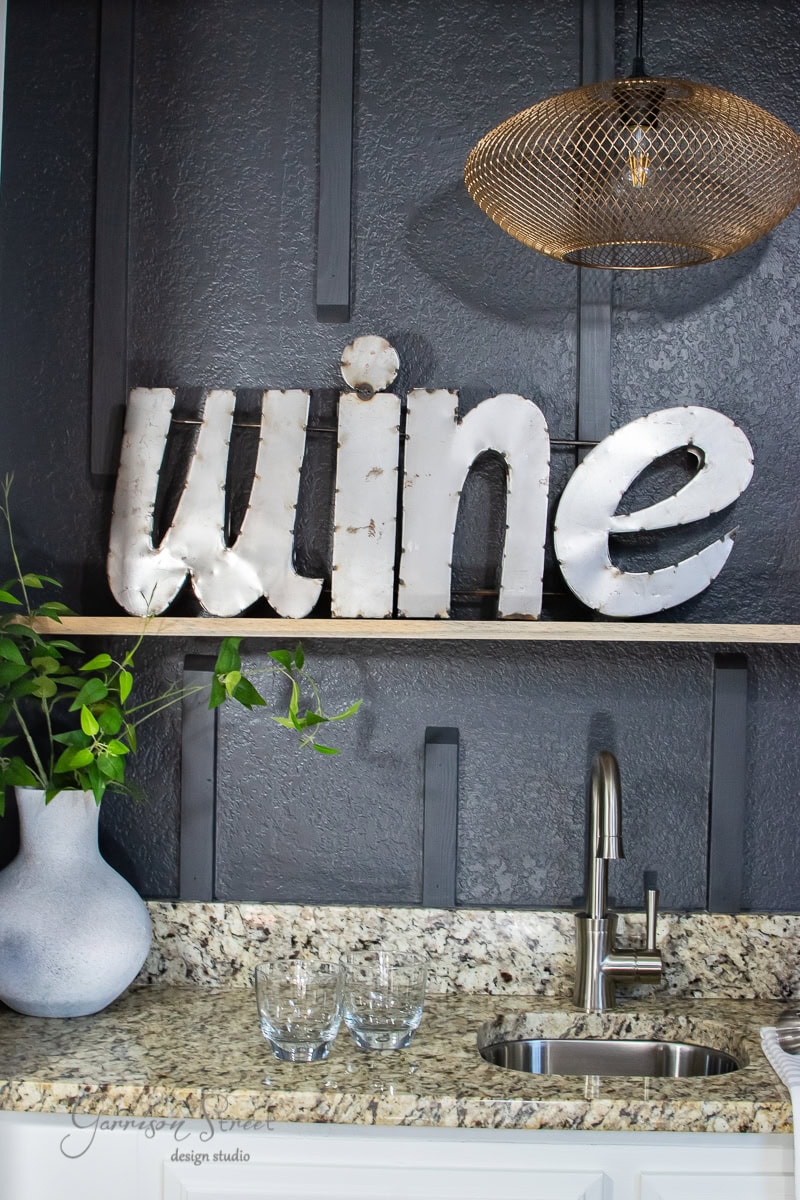A modern kitchen counter showcases a metal "wine" sign on a floating shelf. Below, the wet bar area features a granite surface with a sink, several glass cups, and a white vase with green branches. The backdrop is a textured dark wall, illuminated by a wire mesh pendant light above.