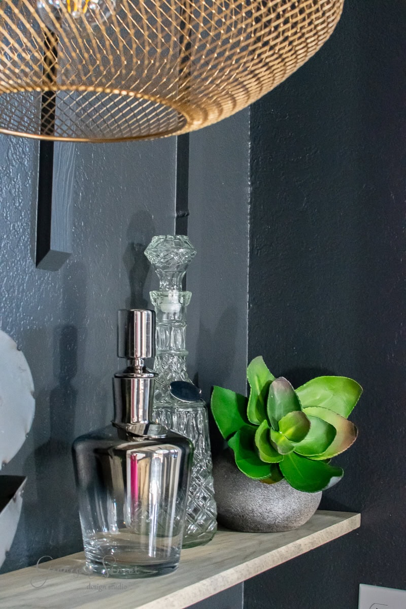 A stylish floating shelf holds two elegant glass decanters and a lush green succulent in a gray pot. Above, a woven rattan light fixture casts a warm glow against the dark wall, perfect for creating a cozy and modern bar ambiance.