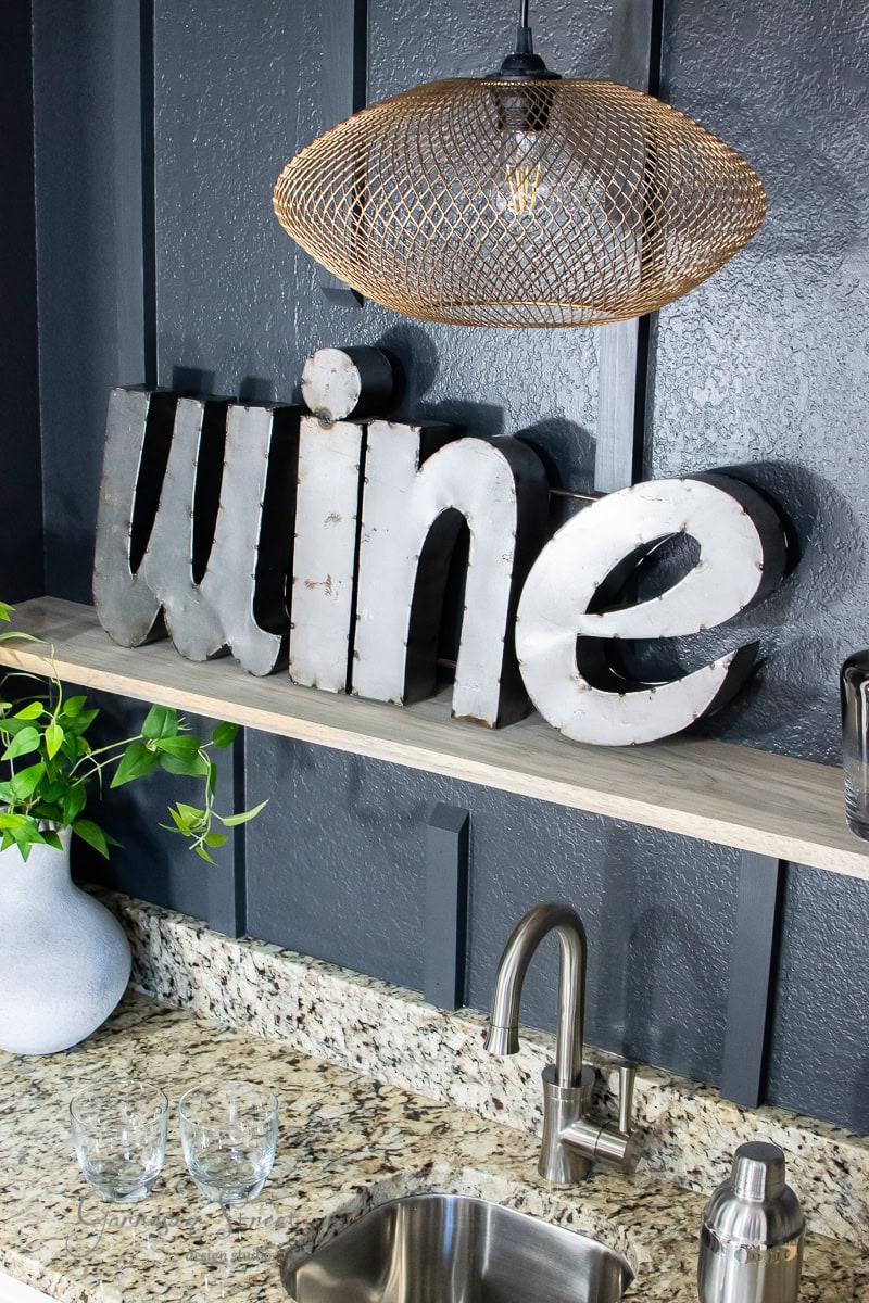A modern kitchen features a metallic "wine" sign on a wooden floating shelf. Below the sign is a granite countertop with a small wet bar sink and two empty glasses. A potted plant sits on the left, and a textured pendant light hangs above.