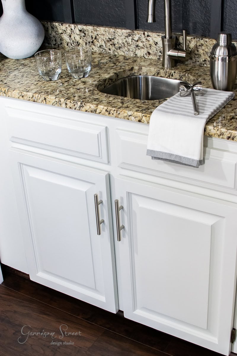 A modern kitchen area featuring a polished granite countertop, stainless steel sink, and shiny faucet. A striped kitchen towel is draped over the sink. Below are white cabinets with metal handles. Two drinking glasses and a metallic shaker sit atop the counter by a wet bar setup.
