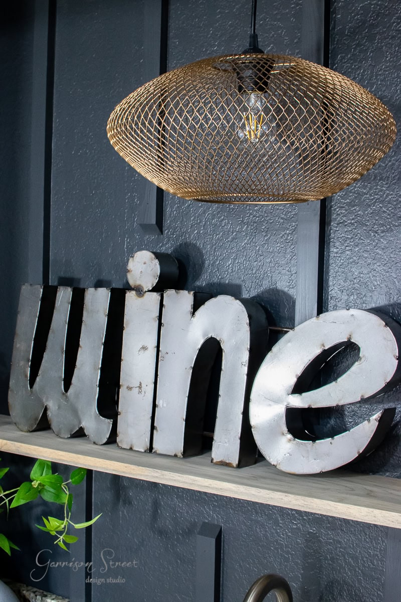 A decorative metal sign spelling "wine" sits on a wooden floating shelf against a dark gray wall, bringing a touch of charm to this cozy wet bar. Above it hangs a gold wire pendant light, while a small plant is partly visible on the left side.