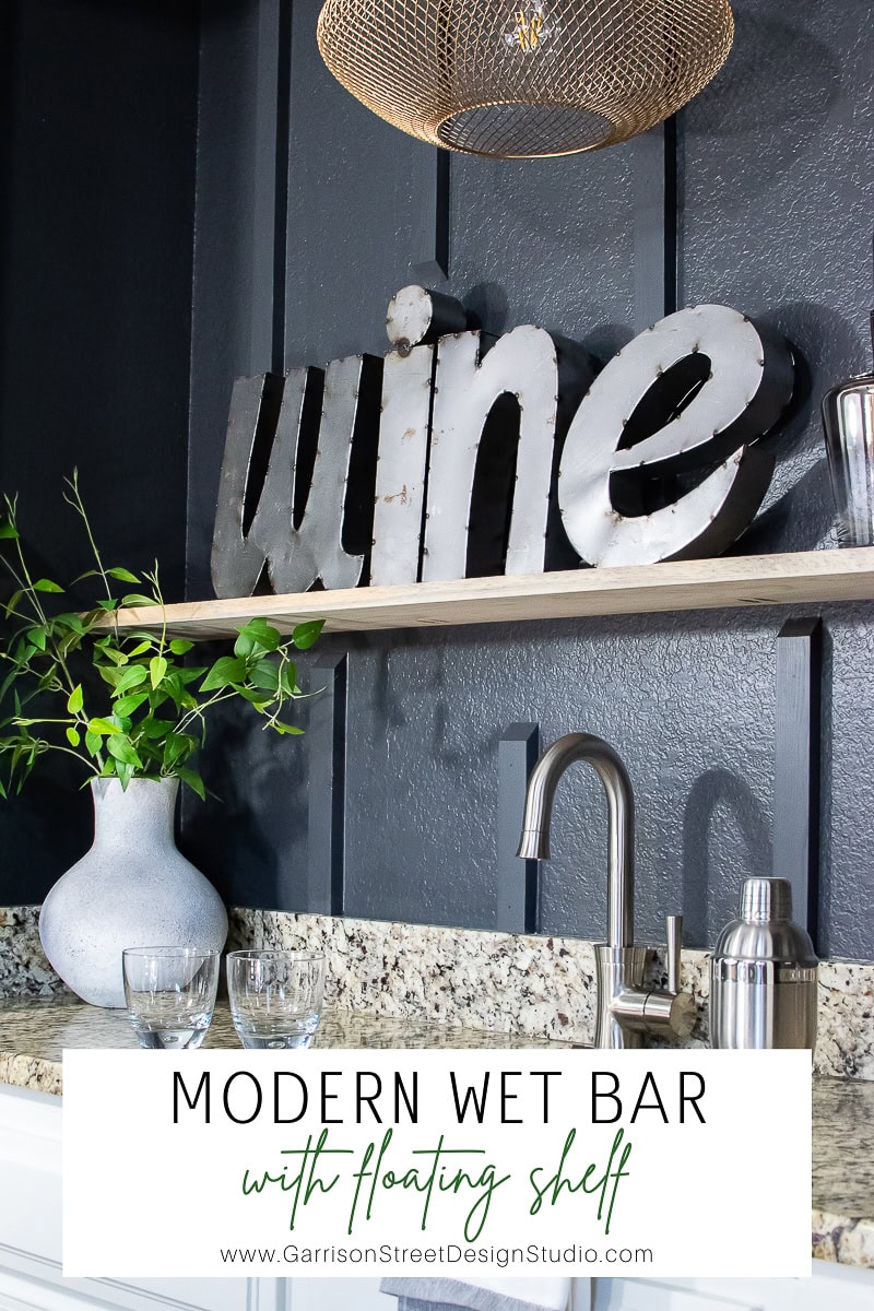 This sleek wet bar boasts a floating shelf with a bold metallic "wine" sign. Complete with a sink, cocktail shaker, glasses, and a vase of green leaves, it sits elegantly on a granite countertop against the dark paneled wall.