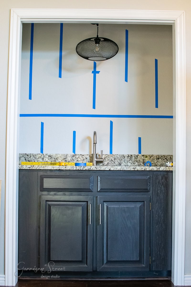 A small kitchen area features dark wood cabinets and a granite countertop. Blue painter's tape on the wall above the sink hints at a planned design, possibly for a floating shelf. A black pendant light adds charm to the space.
