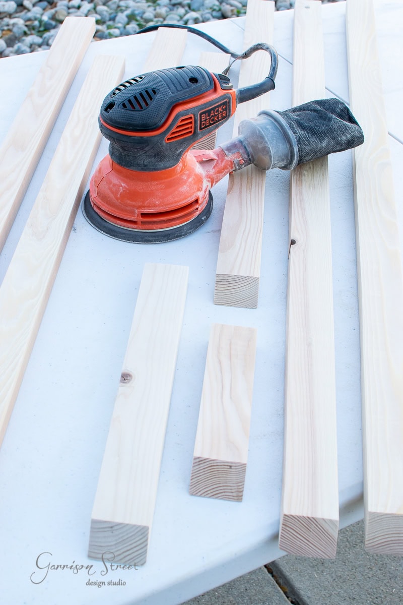 An orange and black orbital sander sits on a white table, surrounded by various pieces of light-colored wood for a floating shelf project. The sander has a dust collection bag attached. Stones are visible in the background, adding contrast to the workshop scene.
