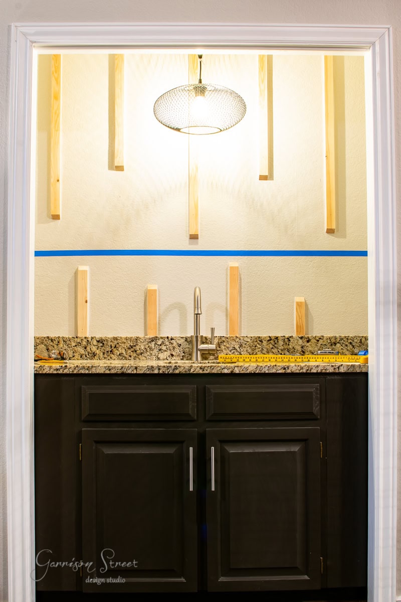 A modern kitchen nook features a granite countertop and sleek faucet set against a beige wall with vertical wooden slats. A floating shelf adds practical elegance above the wet bar, while a wire mesh pendant light illuminates the space. Dark cabinetry provides contrast below the countertop.