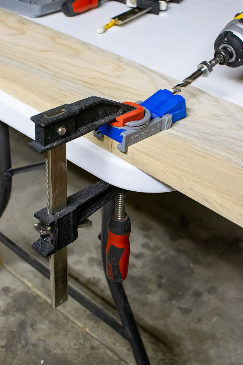 A workbench with a wooden board clamped to the edge resembles a busy bar countertop. A power drill with a bit is inserted into a blue pocket hole jig, and a metal clamp holds the board steady as if crafting the perfect floating shelf. Tools are scattered on the table in organized chaos.
