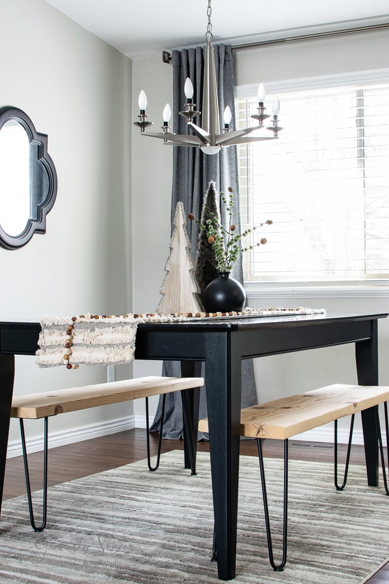 A modern dining room, reminiscent of a 2020 Christmas house tour, features a black table with bench seating. A black vase with flowers sits on the table, alongside small decorative trees. A chandelier hangs above, while curtains frame the window and a geometric mirror adorns the wall.