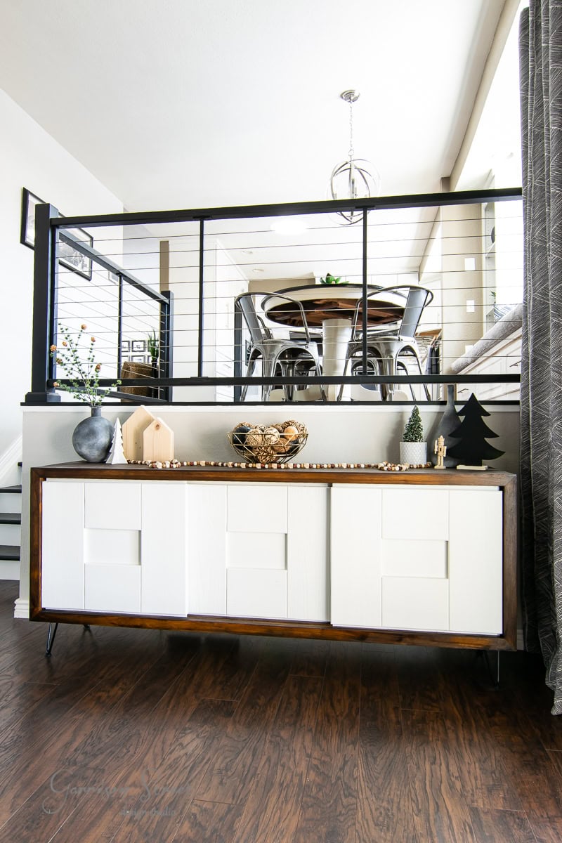 Modern interior with a white sideboard on dark wooden floors. Decorative items, like pine trees, hint at a Christmas house tour vibe. A metal railing separates the area from a dining space featuring chairs and a round table. Pendant light hangs from the ceiling, adding to the 2020 charm.