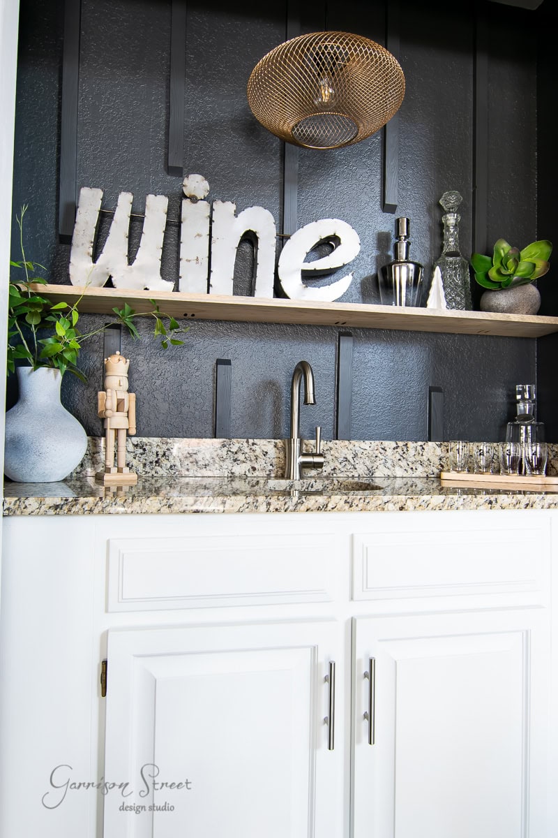 Home bar with white cabinets and a granite countertop perfect for a cozy Christmas house tour. The black wall features a wooden shelf adorned with festive decorative items, including a large "wine" sign, metal shaker, and plants. A brass pendant light adds charm to the 2020 vibe.