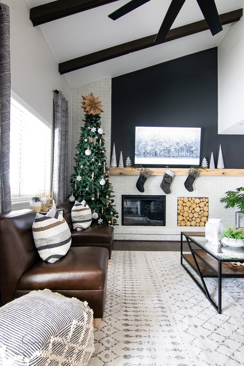 A modern living room, perfect for a 2020 Christmas house tour, features a tall tree adorned with ornaments and a star. Stockings hang on the mantel above the fireplace. The room boasts a cozy sofa, patterned pillows, and a coffee table resting on a textured rug.