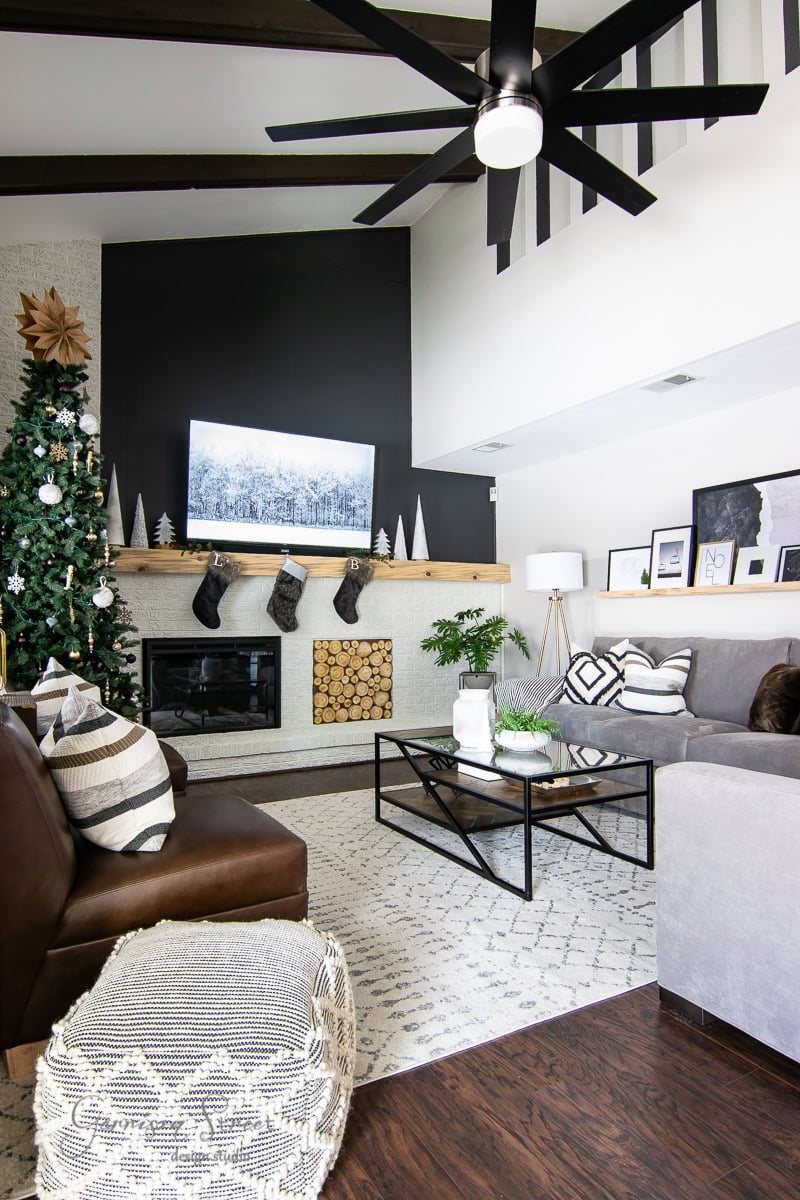 Experience a 2020 Christmas house tour in this modern living room, where a beautifully decorated Christmas tree stands beside a cozy fireplace adorned with festive stockings. A large black-and-white photo graces the wall above the mantel's wood stack, while a gray sofa and dark brown chair complete the warm seating area beneath the sleek black ceiling fan.