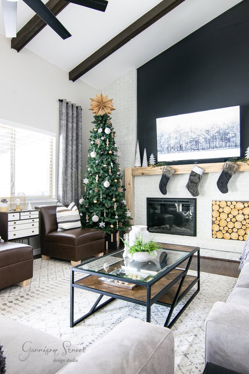 A modern living room, perfect for a 2020 Christmas house tour, features a tall tree adorned with ornaments beside a cozy fireplace. Stockings dangle above the firewood-filled hearth. The space boasts comfortable seating, a stylish coffee table, and large windows with elegant curtains.