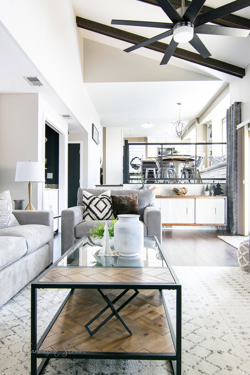 Experience a modern living room featuring a sleek gray sofa and a glass coffee table adorned with geometric decor. A black ceiling fan and exposed beams add character, while the bright, airy space welcomes you to continue your 2020 Christmas house tour through the dining area in the background.