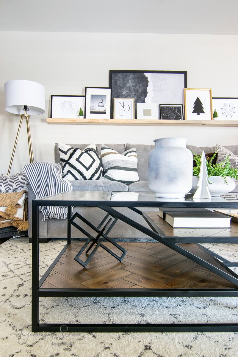 Experience a modern living room perfect for any 2020 Christmas house tour. It features a gray sofa with geometric patterned cushions, a low black metal and wood coffee table, and decorative items on a shelf above. A floor lamp stands on the left, illuminating the textured rug below.