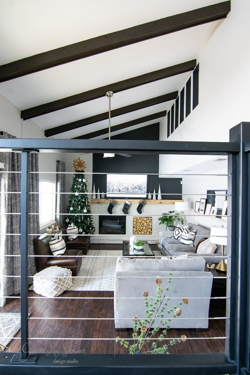 Experience a modern living room with high, beamed ceilings and a cozy fireplace, perfect for your Christmas house tour. A decorated tree stands by the large window, alongside grey sofas and a coffee table on a patterned rug. Shelves boast decorative items and framed pictures, capturing 2020 memories.