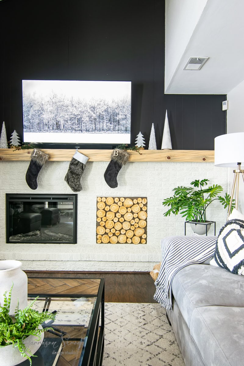 In this 2020 Christmas house tour, a modern living room features a white brick fireplace with a wooden mantel adorned with stockings and small white trees. Above, a landscape painting hangs gracefully. A cozy gray sofa sits to the right, enhanced by cushions and a striped blanket.