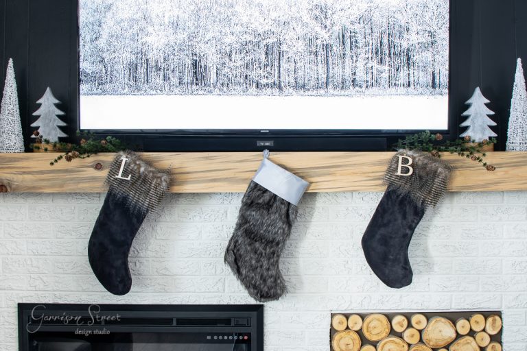 Step into a cozy 2020 Christmas house tour, where a rustic wooden mantel graces the charming fireplace scene. Stockings with initials "L" and "B" dangle beneath a snowy forest landscape painting, while decorative trees and logs add festive warmth to the inviting space.