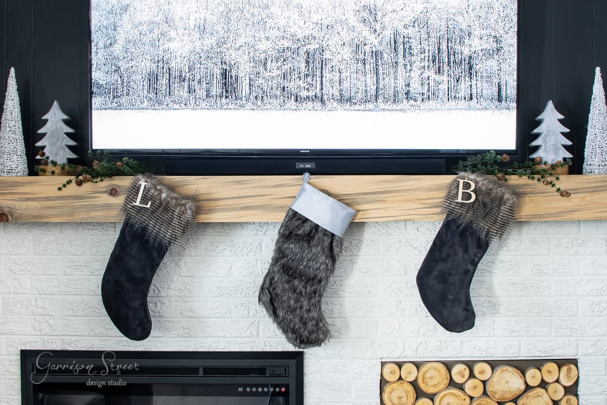 Step into a cozy 2020 Christmas house tour, where a rustic wooden mantel graces the charming fireplace scene. Stockings with initials "L" and "B" dangle beneath a snowy forest landscape painting, while decorative trees and logs add festive warmth to the inviting space.