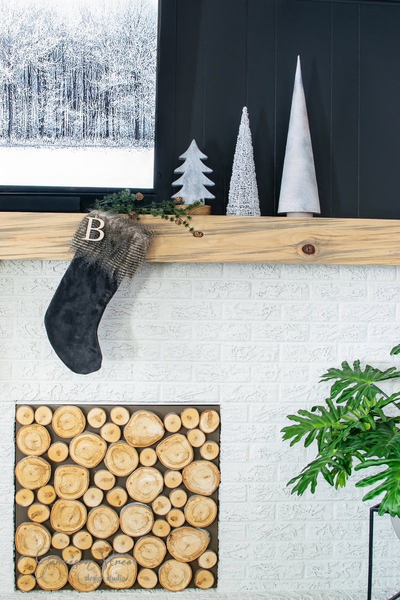 A fireplace mantel with minimalist decor sets the scene for a 2020 Christmas house tour: three cone-shaped trees and a black stocking with a "B" hanging from it. Below, the fireplace is filled with stacked logs, while a green plant adds a touch of nature to the right.